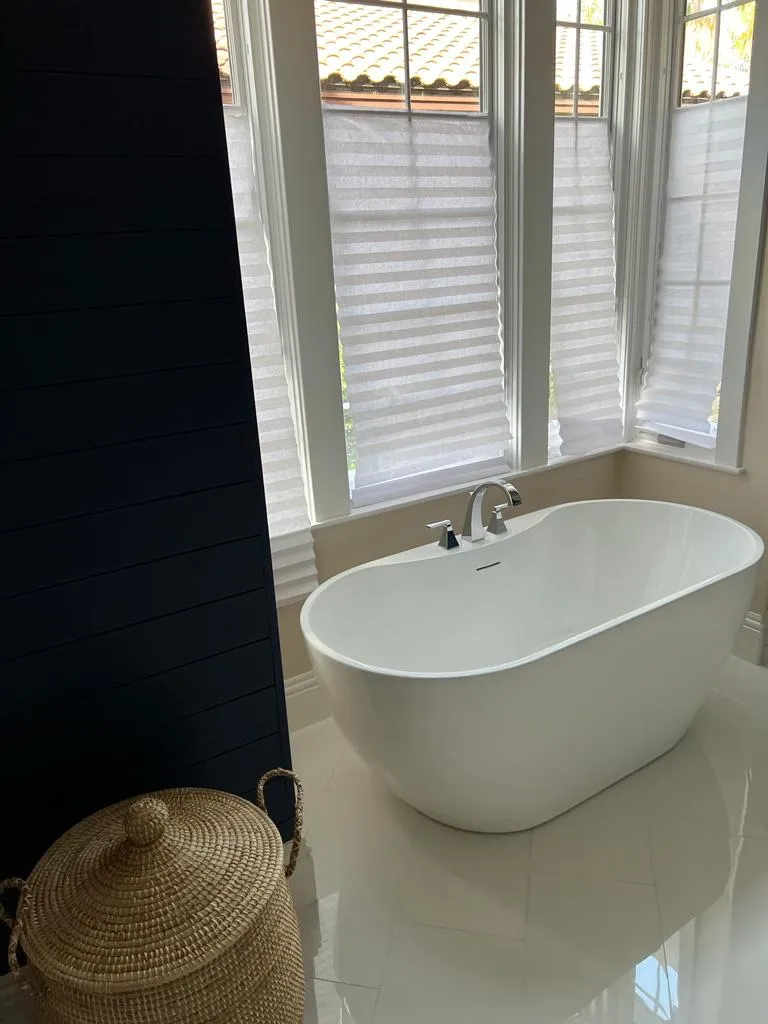 Serene bathroom design with white freestanding bathtub positioned by large windows with plantation shutters and woven basket accent